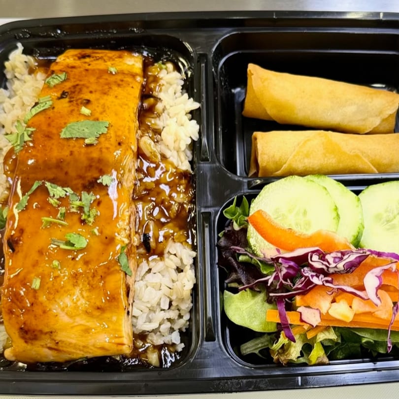 Teriyaki salmon with rice, two spring rolls, and a side salad in a black bento box.
