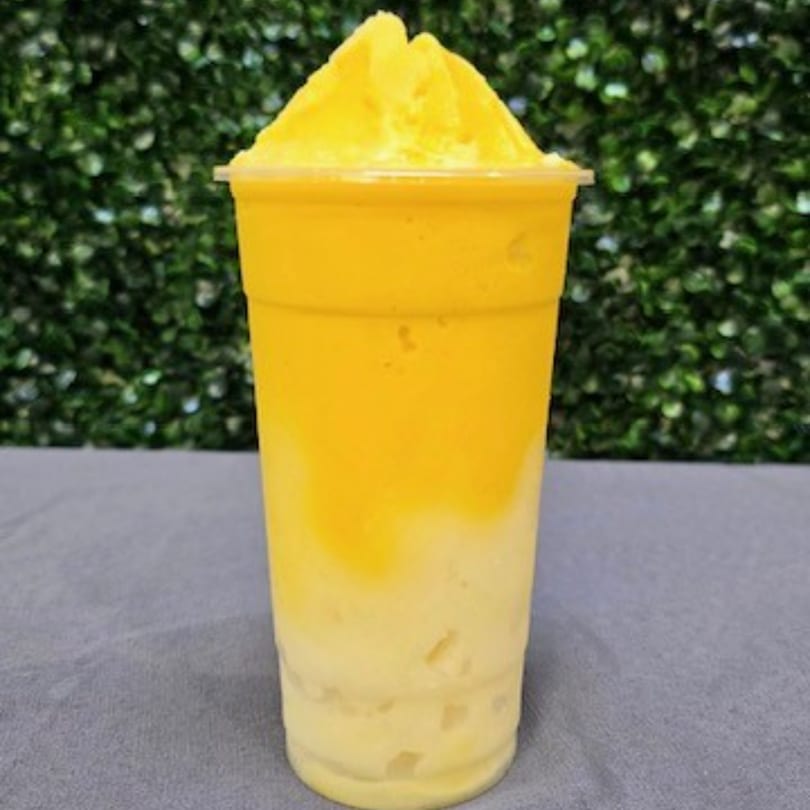 A two-toned mango smoothie, yellow on top and lighter yellow on the bottom, against a green leafy background.